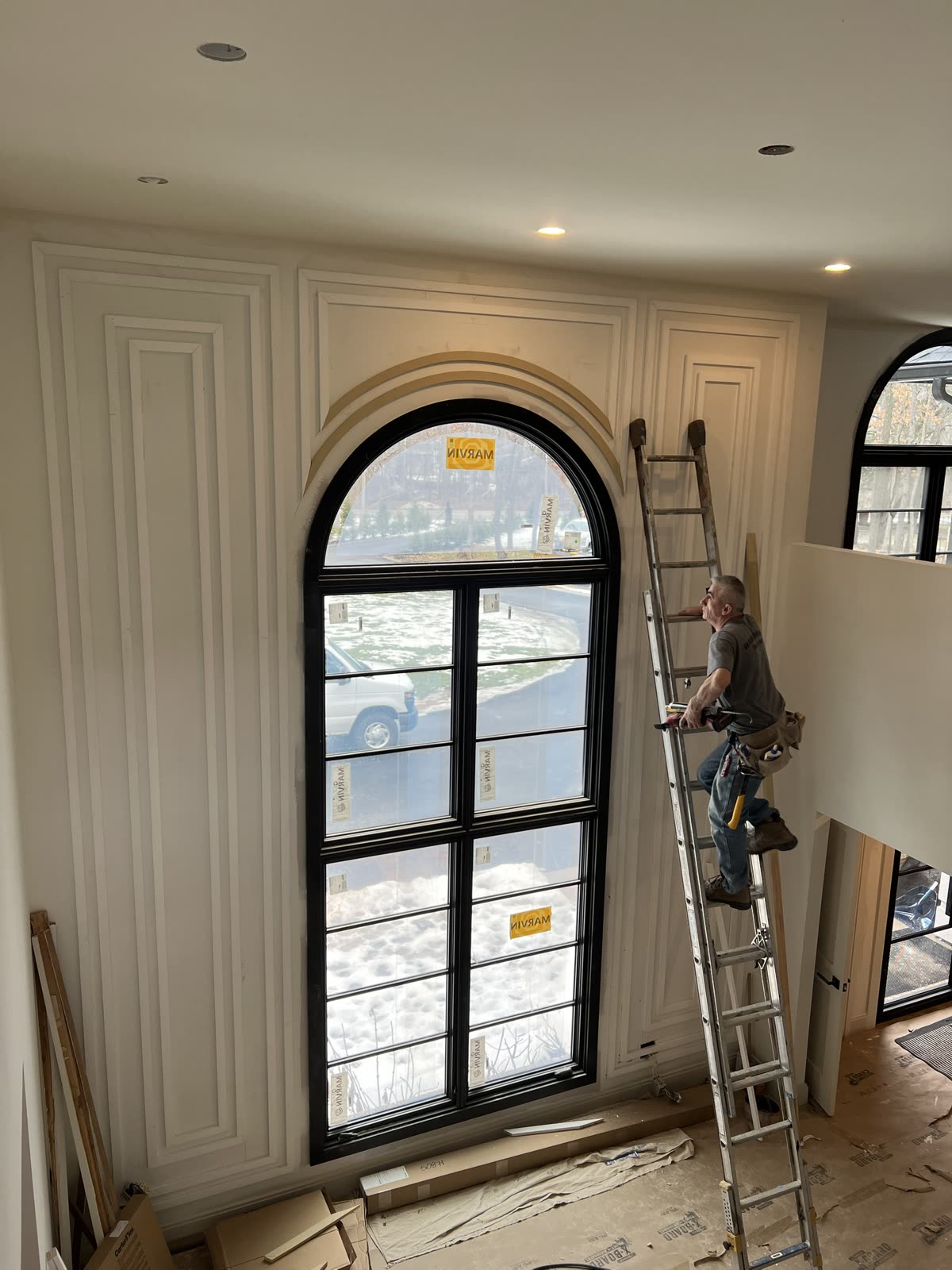 Two-story foyer before paint: picture-frame molding on both sides of a tall black arched window, a curved arch casing being fitted over it, and a carpenter on a ladder.