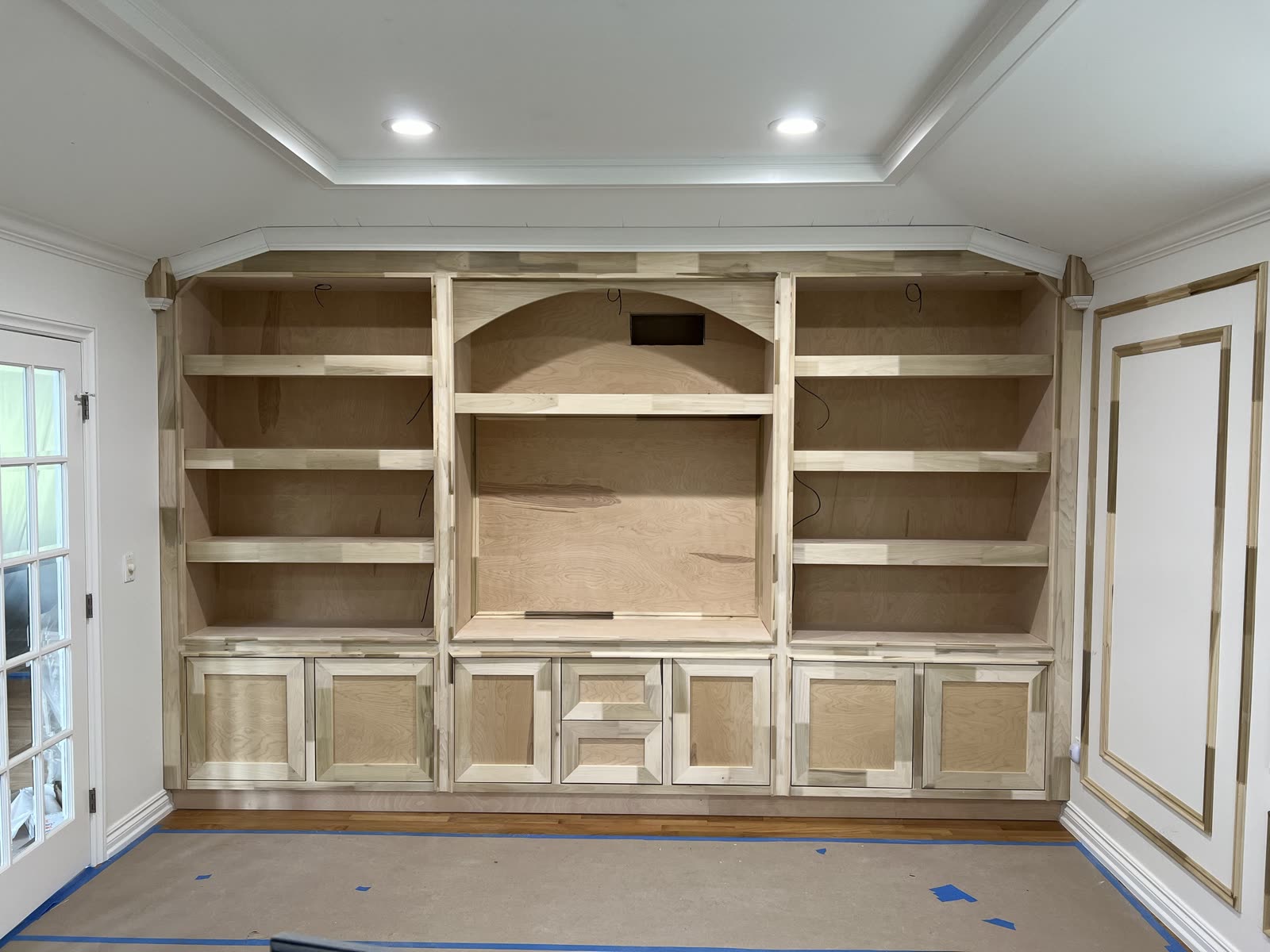 Wall-to-wall built-in before paint, raw wood: open shelves on both sides of an arched center niche, shaker doors and drawers along the bottom, crown at the ceiling.
