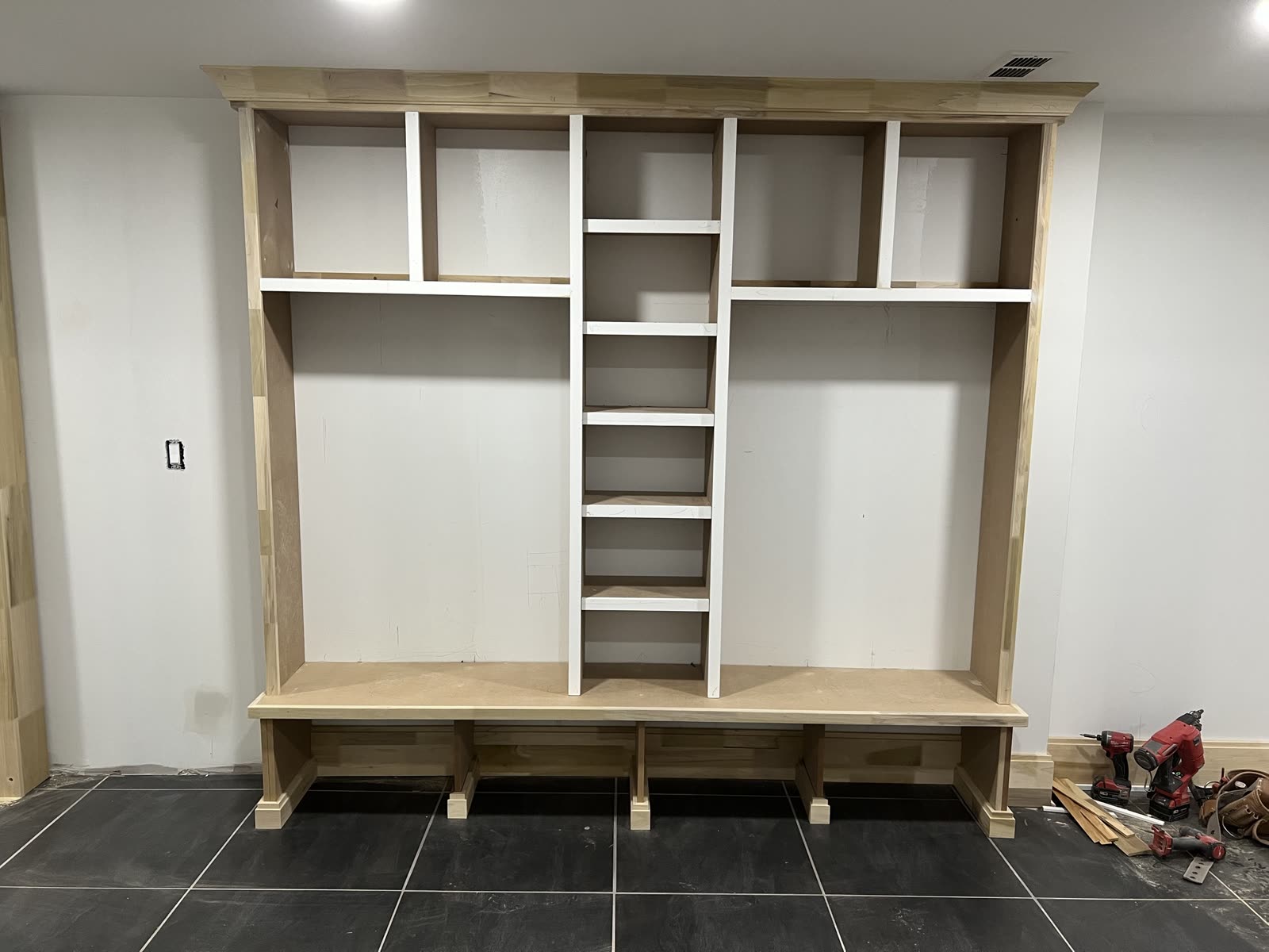 Built-in mudroom bench and locker unit before paint: crown molding across the top, a narrow ladder of shelves down the center, a bench seat on plinth feet, raw wood and MDF on a black tile floor.