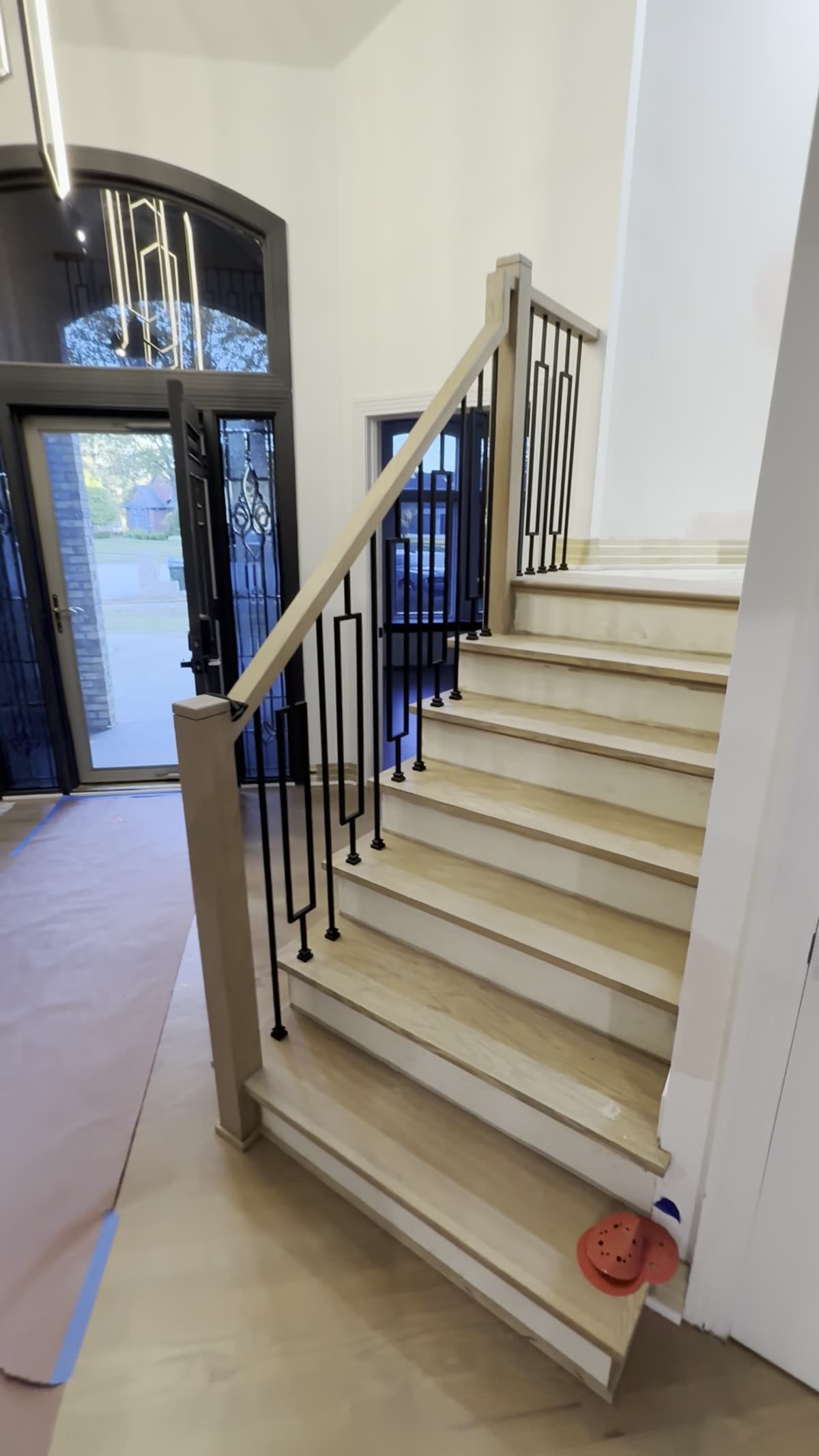 Staircase before finish: light wood treads and risers, a square newel post and handrail, and black iron balusters with rectangular loops, with the front door behind.